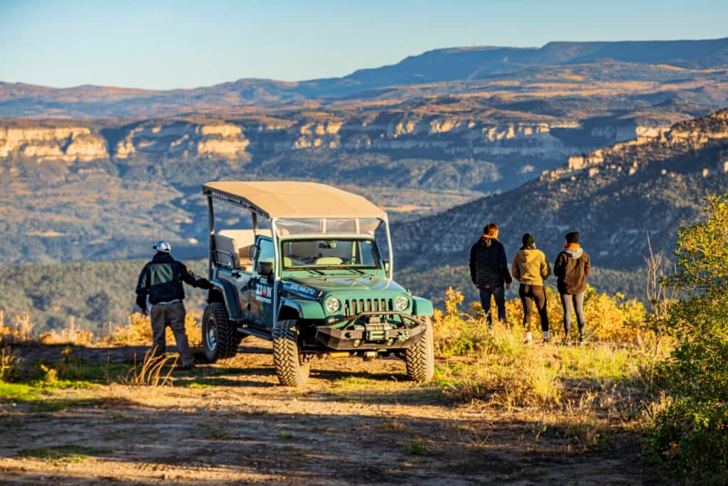 A group of four people stands near a jeep, overlooking a scenic mountain landscape.