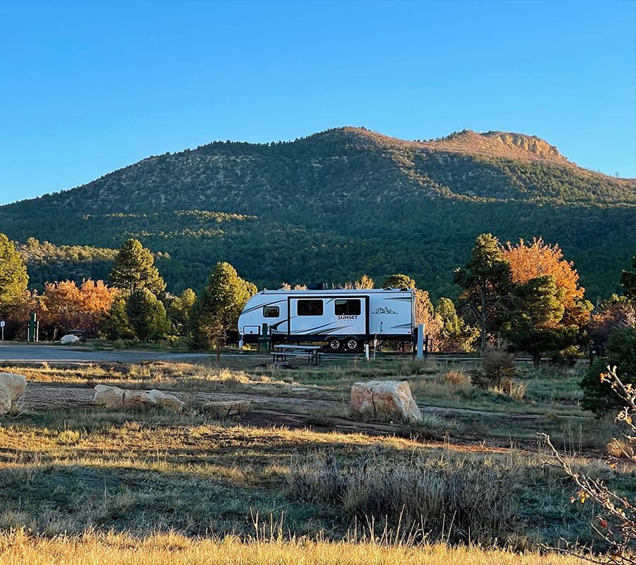 A white RV parked near a mountain surrounded by trees under a clear blue sky.
