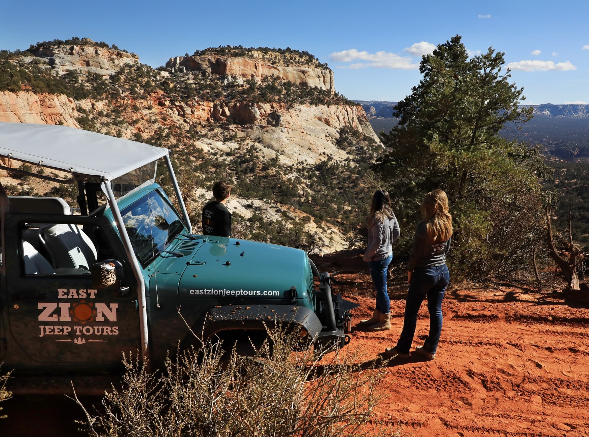 Three people stand beside a green jeep, overlooking a scenic canyon landscape.