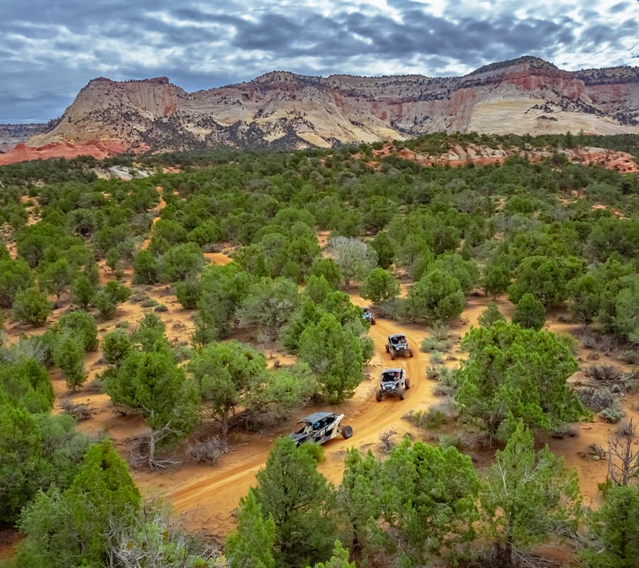 Off-road vehicles traverse a dirt path through a lush green landscape with rocky cliffs in the background.
