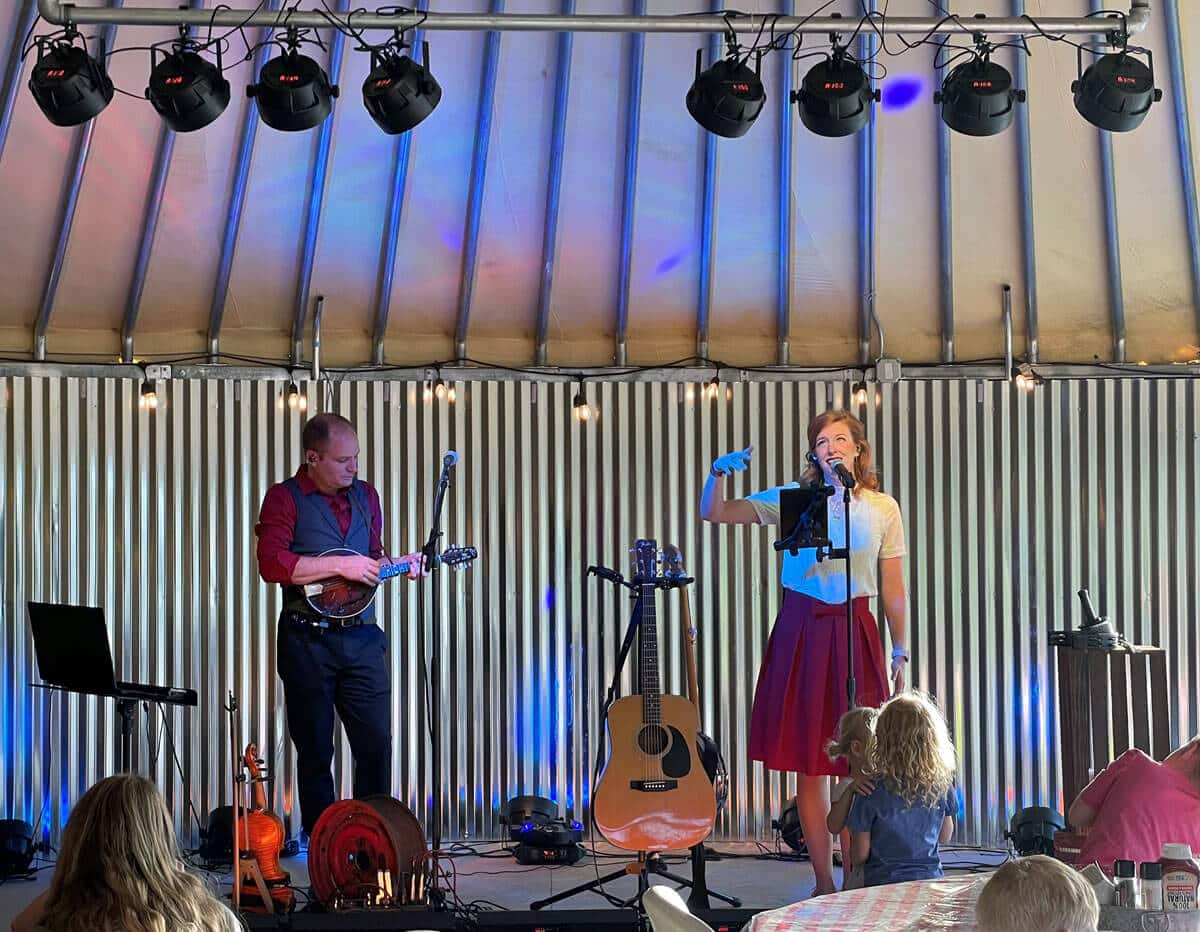 A woman sings into a microphone while a man plays a mandolin on stage, with an audience in the foreground.