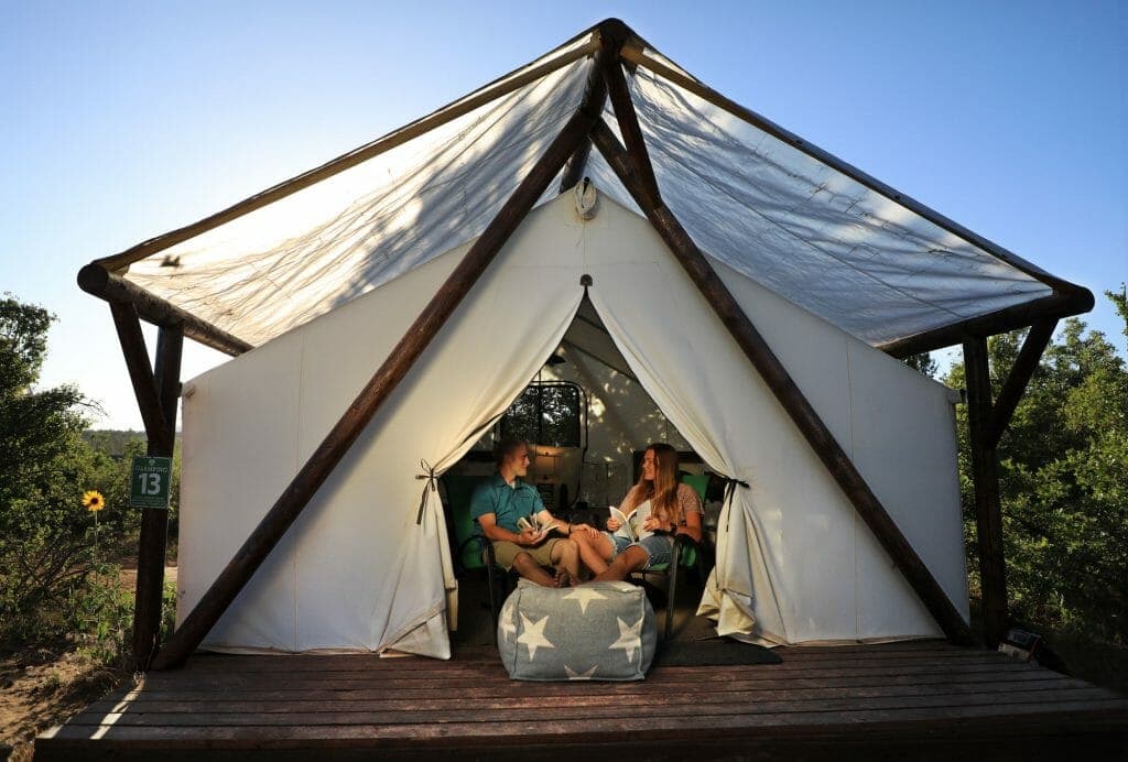 A couple relaxes inside a tent with wooden beams, enjoying the outdoors on a sunny day.