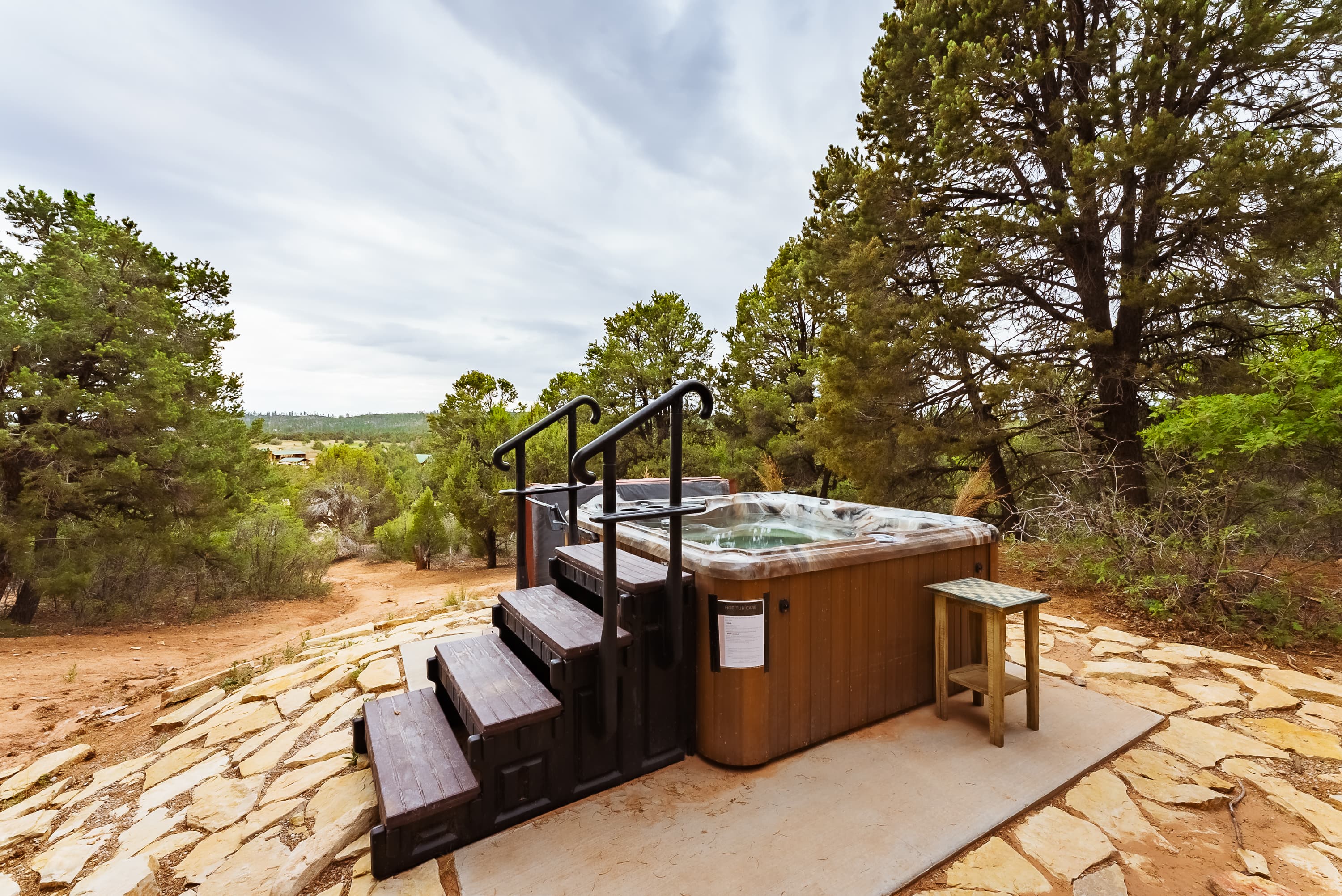 A hot tub with wooden steps is surrounded by trees and rocky terrain.