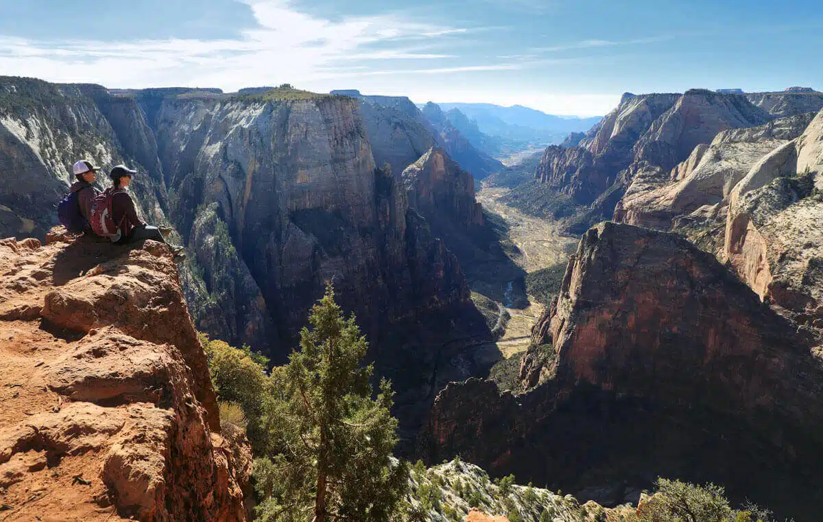 Two hikers sit on a rocky outcrop overlooking a vast canyon landscape.