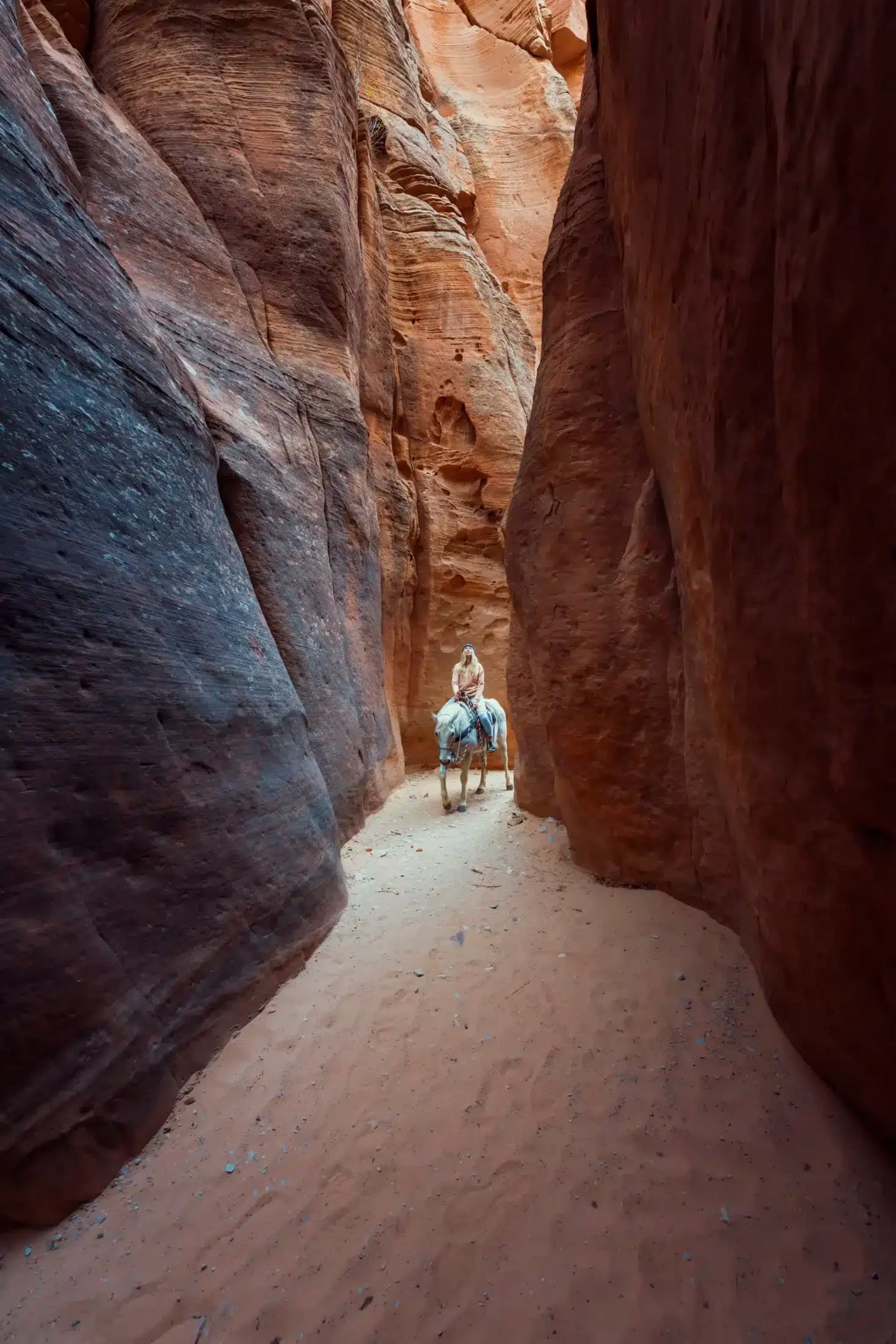 A person rides a horse through a narrow canyon filled with reddish rocks.