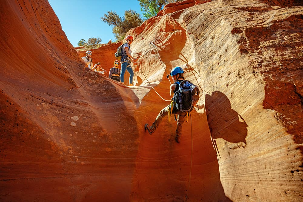 A climber rappels down a red rock canyon while peers watch from above.