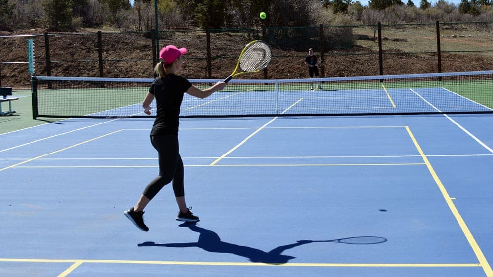A woman in a pink cap hits a tennis ball on an outdoor court while another player stands in the background.