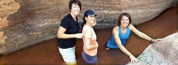 Three women smile while standing in shallow water surrounded by rock formations.