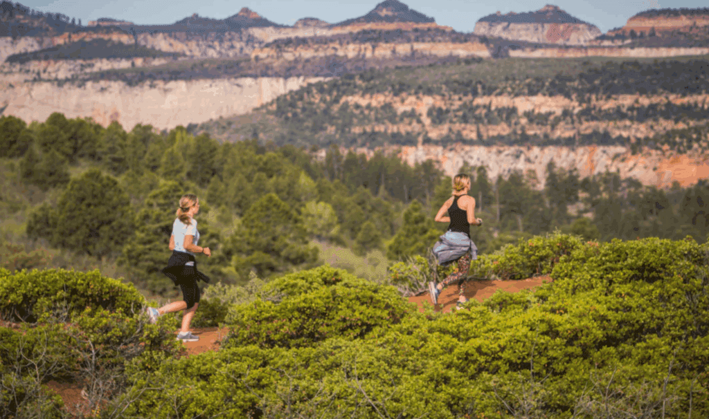 Two women run along a scenic trail surrounded by lush greenery and mountains.