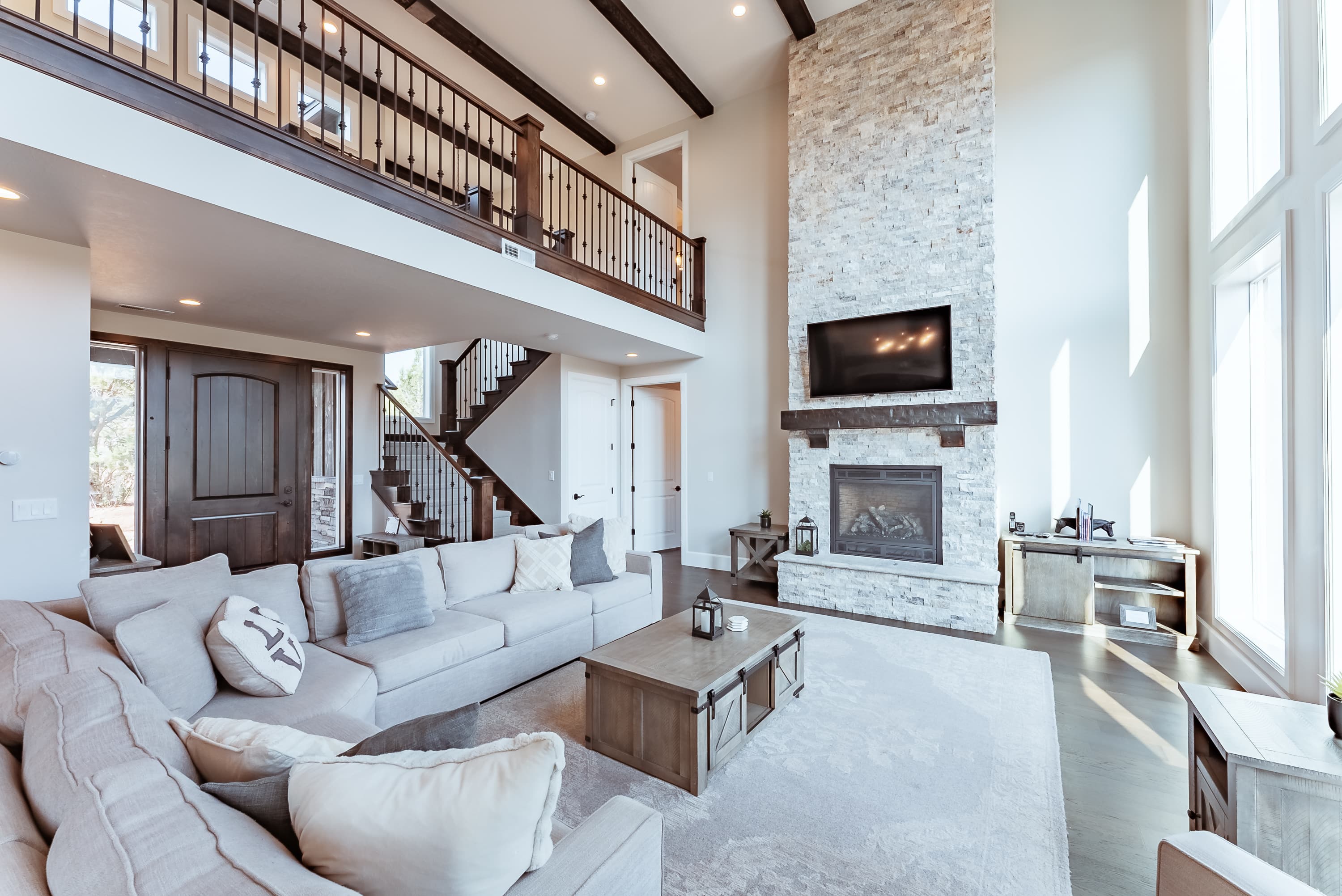 A spacious, modern living room featuring a stone fireplace, large windows, and an open staircase.