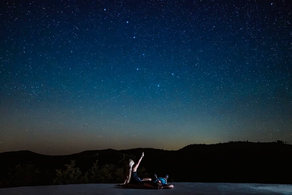 Two people lie on a surface, gazing up at a starry night sky.