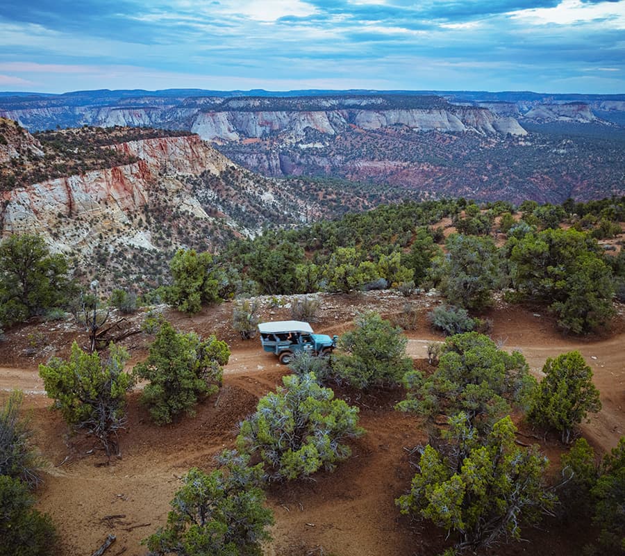 A blue vehicle is parked along a winding dirt road surrounded by greenery, overlooking a rugged canyon landscape under a cloudy sky.