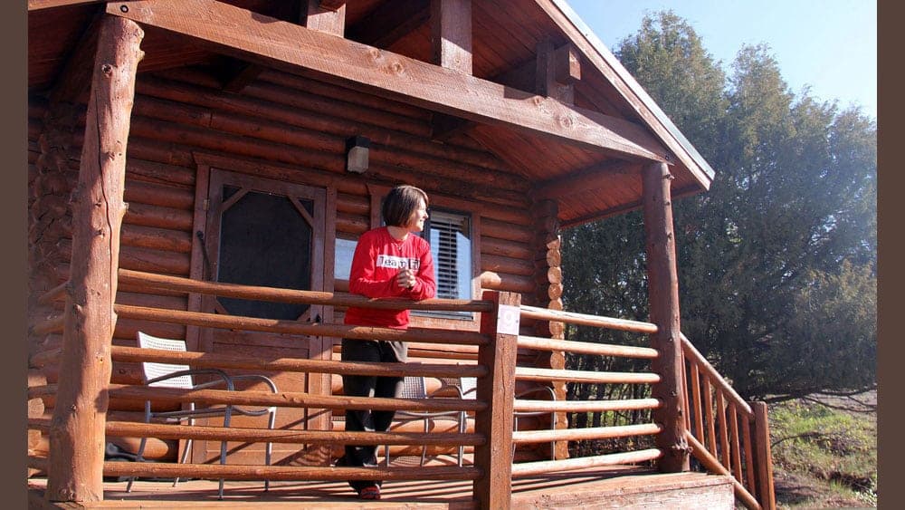 A person stands on the balcony of a log cabin, overlooking a natural landscape.