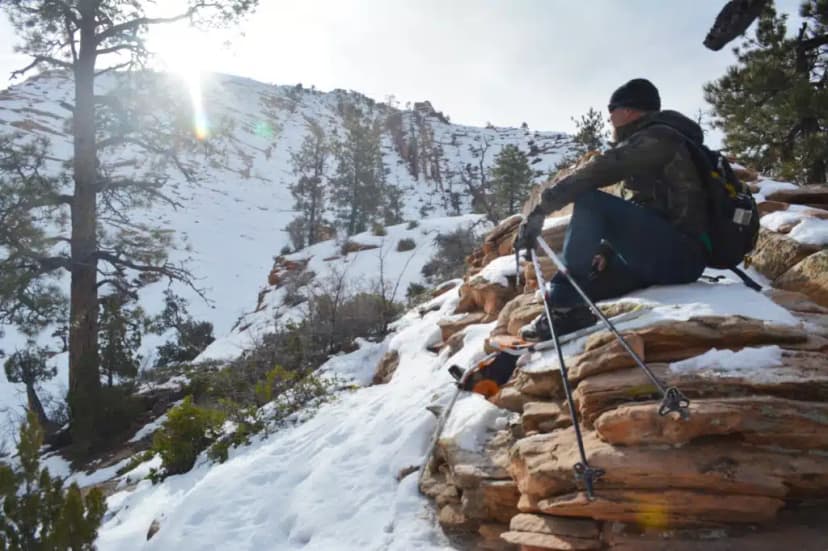 A person sitting on a rocky outcrop covered in snow, resting with hiking poles in a winter landscape.