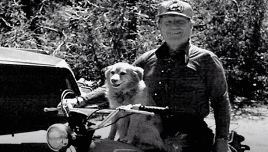 A person in a hat smiles while sitting on a motorcycle next to a dog.