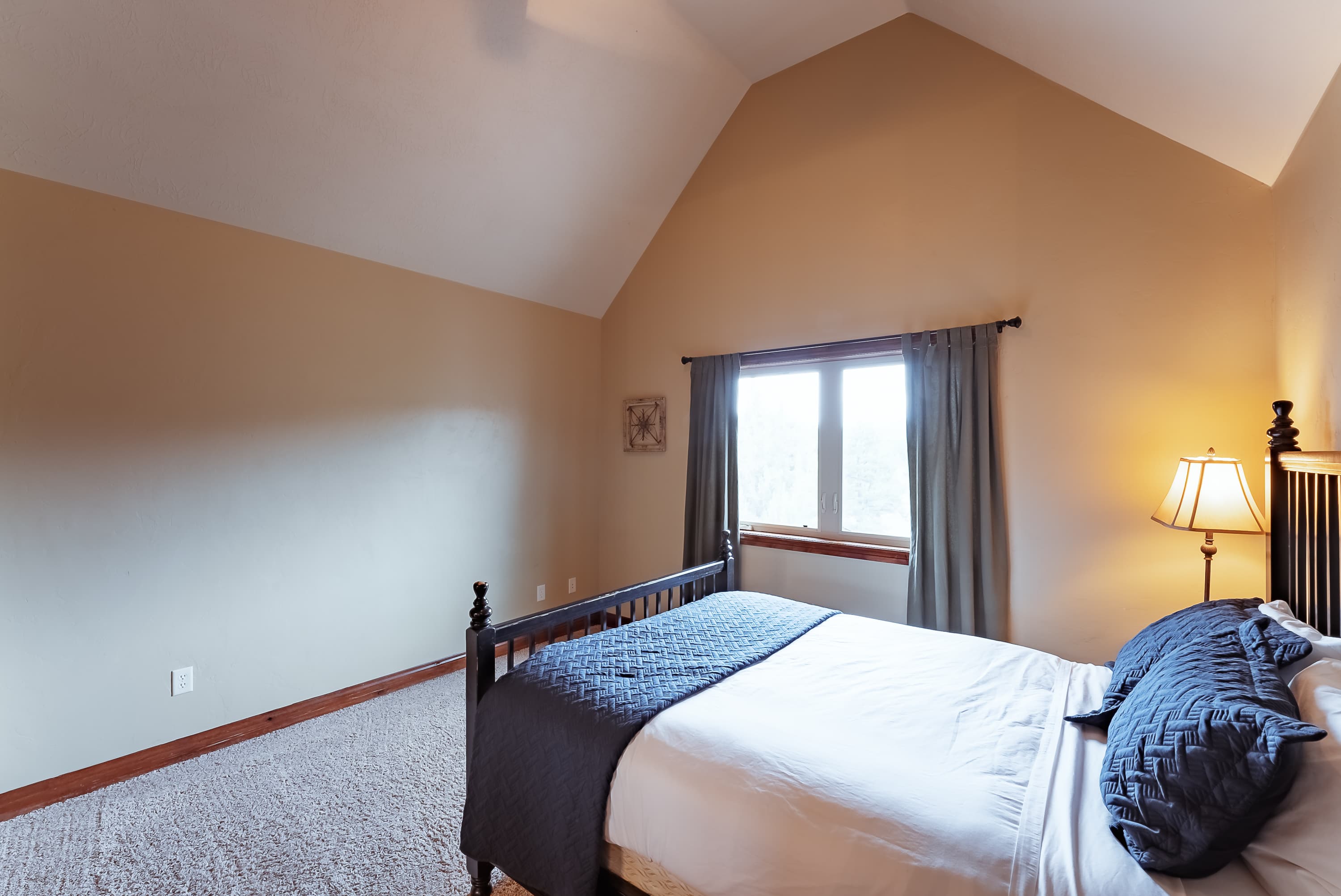 A cozy bedroom featuring a bed with blue accents and a large window beside a lamp.