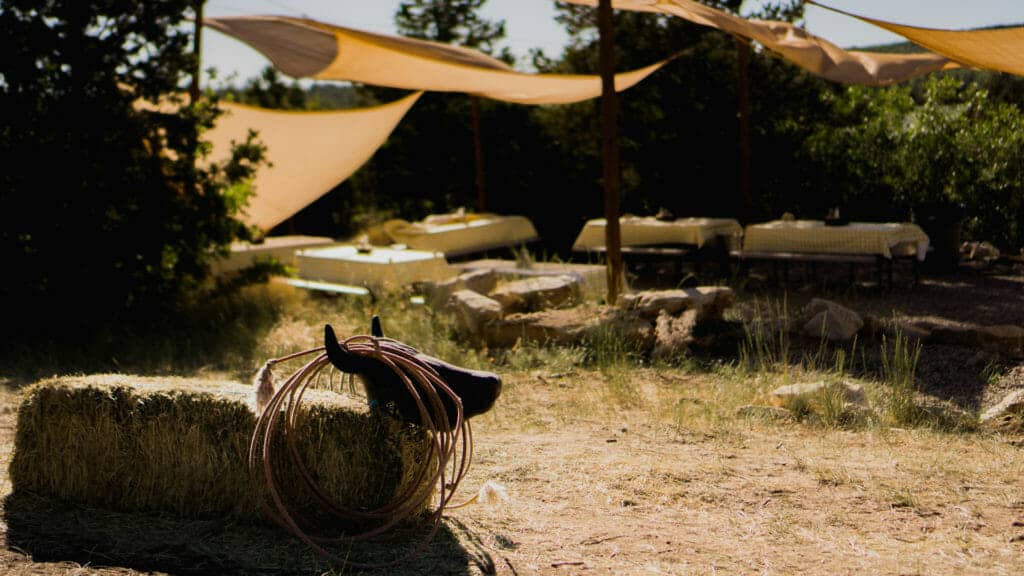 A saddle and rope rest on hay in a rustic outdoor setting with tables under shade sails.