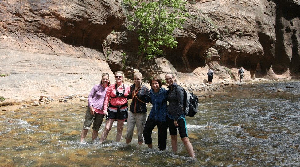 Five women stand together in a shallow stream surrounded by rocky terrain and greenery.