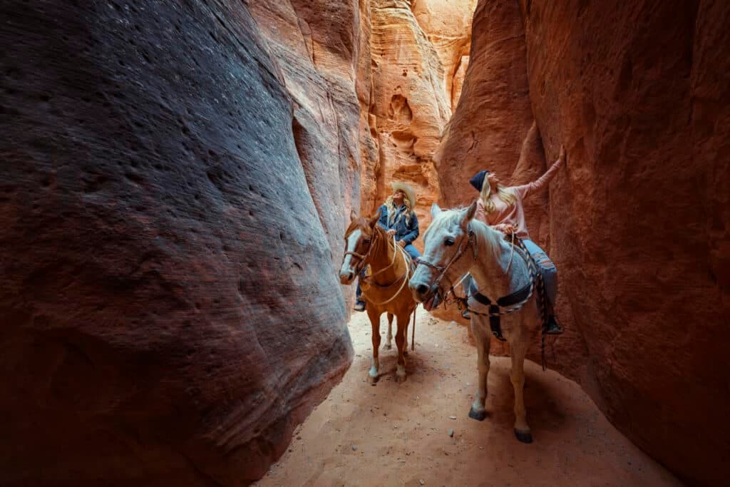 Two people on horseback navigate a narrow canyon.