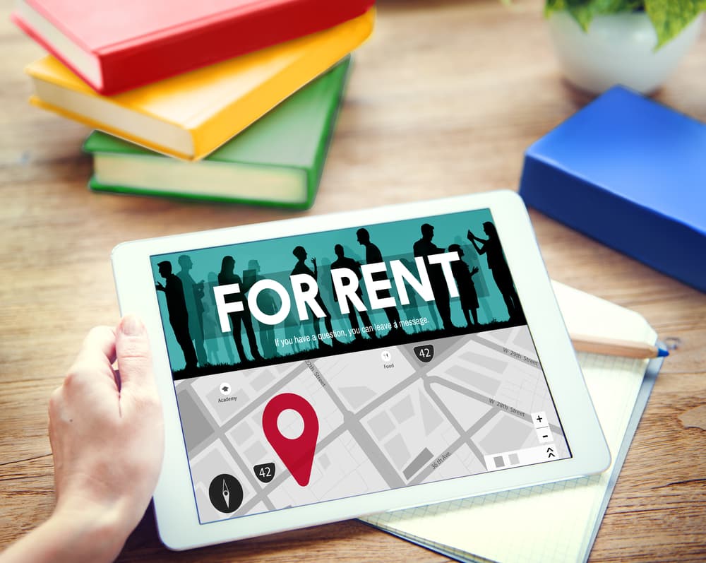 A tablet displays a "For Rent" advertisement with a map and location marker, surrounded by books.
