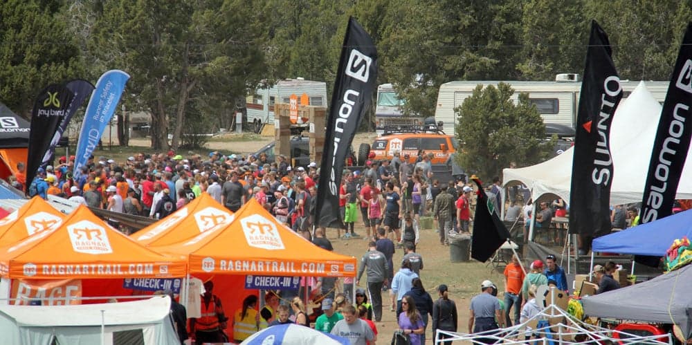 A crowd of participants gathers at a race event with orange and black tents and banners in a park setting.