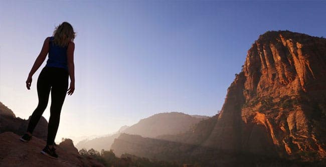 A lone hiker stands on a rocky ledge, gazing at a towering mountain at sunset.