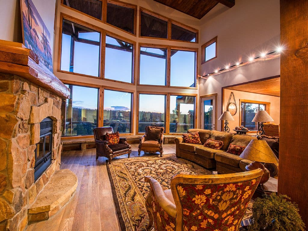 A spacious, warmly decorated living room with large windows offering a scenic view and a stone fireplace.