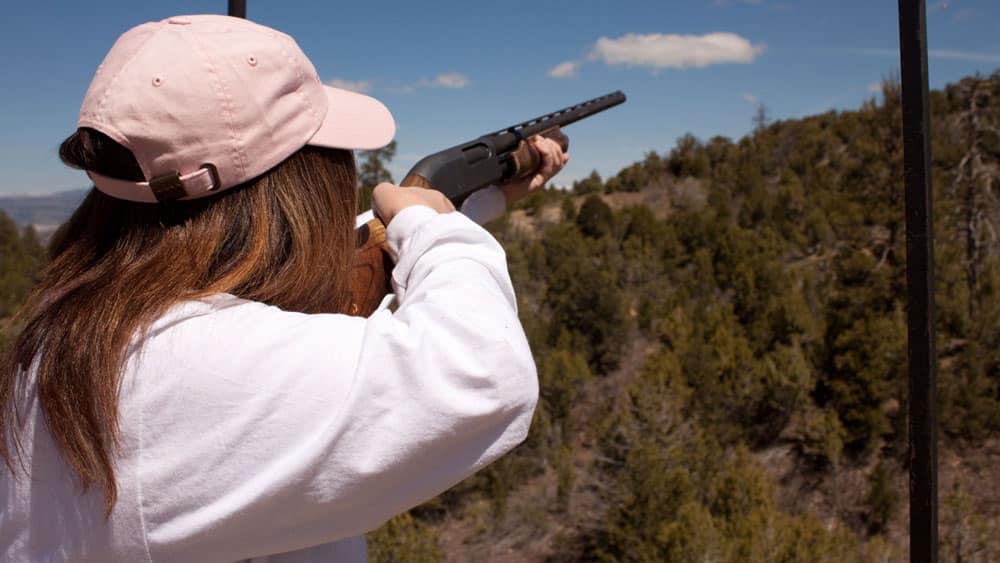 A person in a pink cap aims a shotgun at a distant target in a wooded area.