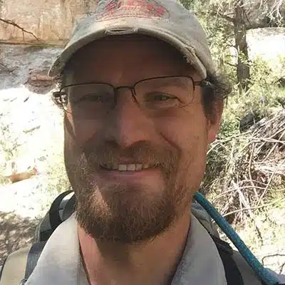 A smiling person with glasses and a cap stands outdoors among trees.