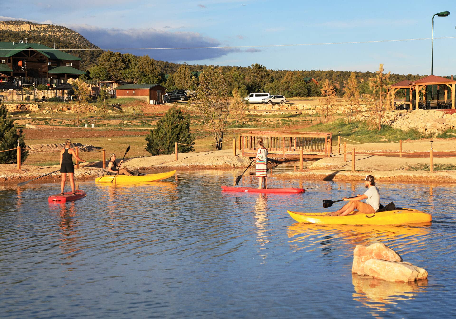 Four people kayak on a calm lake with a scenic backdrop of trees and buildings.