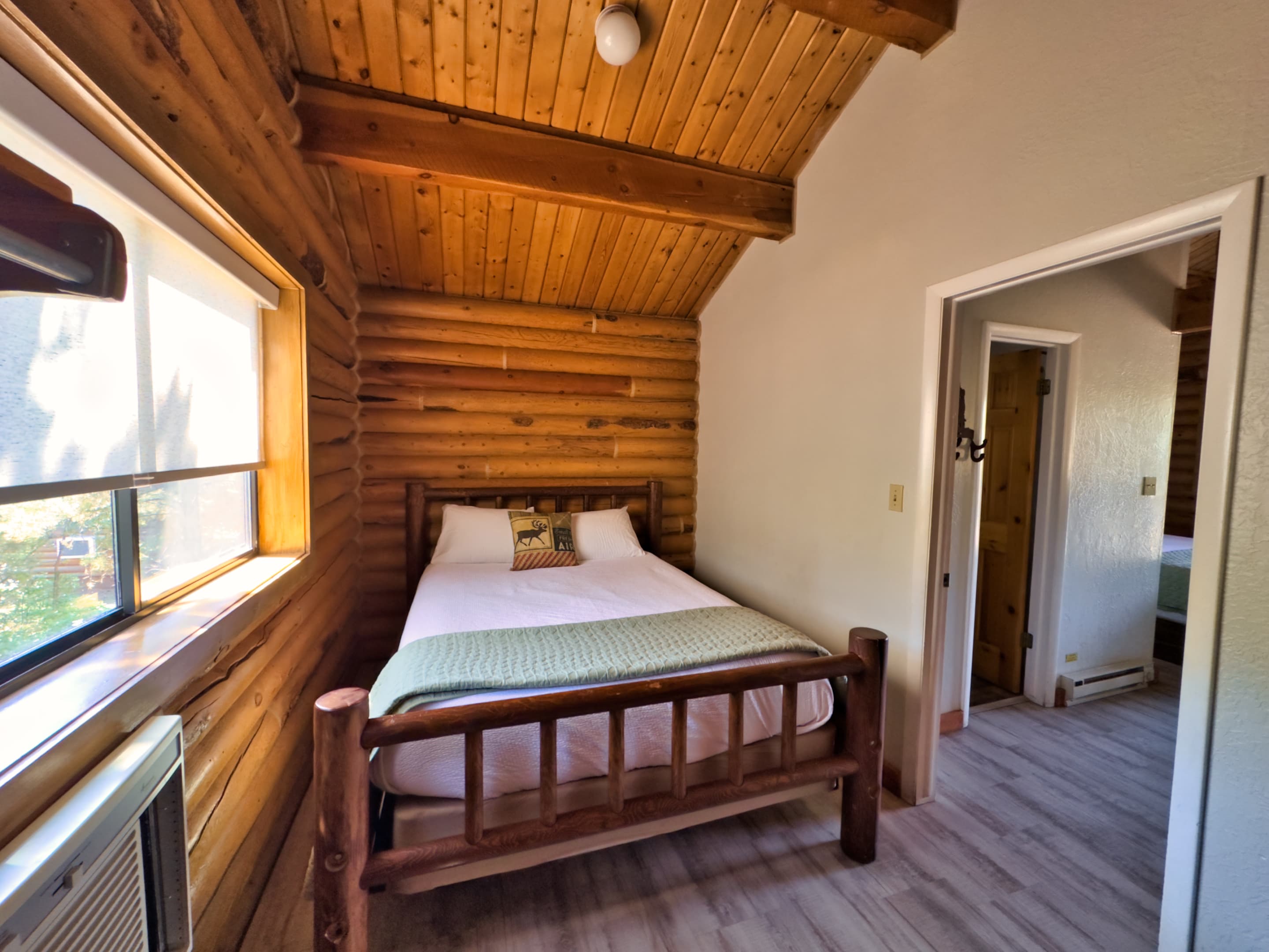 A cozy bedroom with a log-style bed, wooden walls, and natural light from a large window.