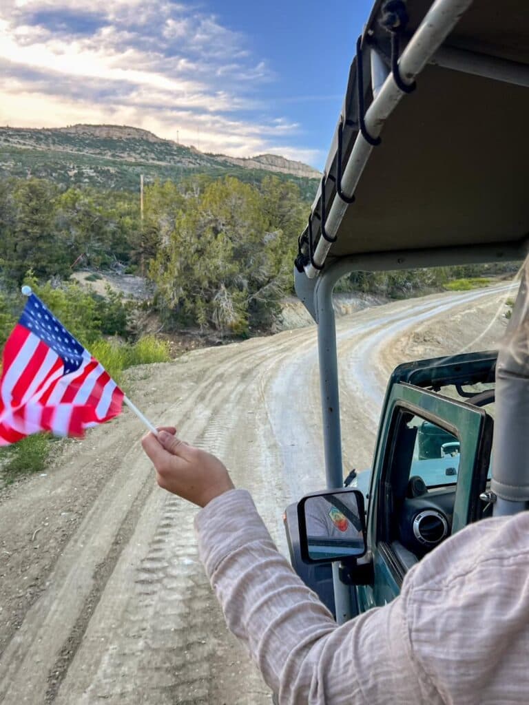 A person in a jeep holds a small American flag while driving along a winding dirt road.