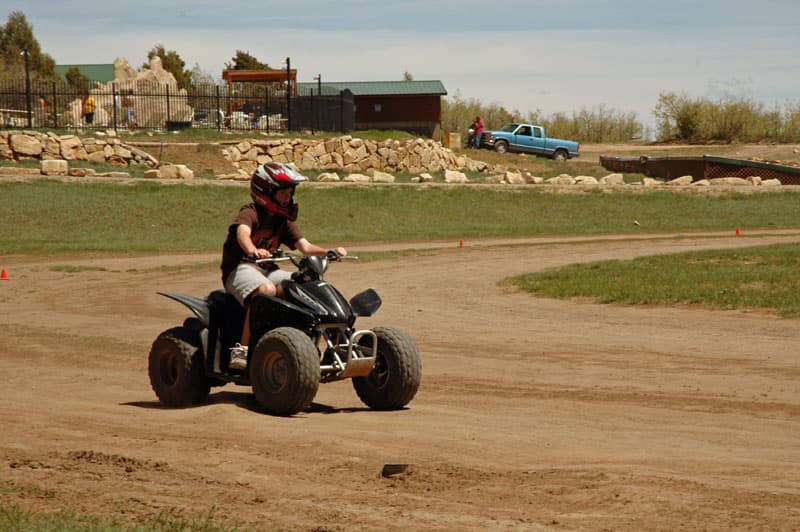A person rides an ATV on a dirt track surrounded by green grass and rocky terrain.