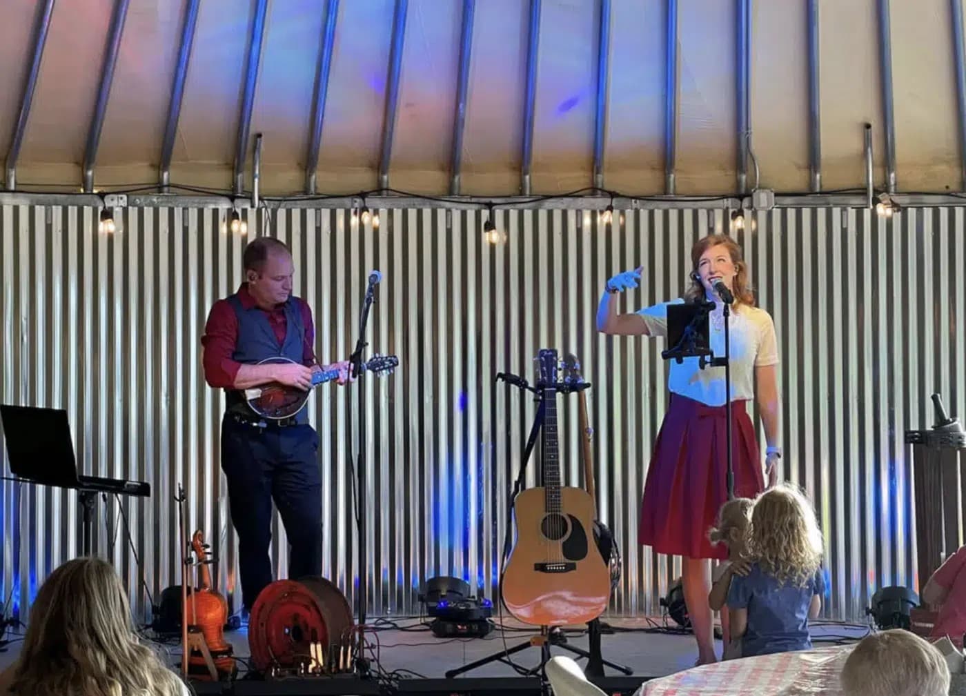 A female singer with a microphone performs on stage alongside a man playing a mandolin, with several musical instruments visible.