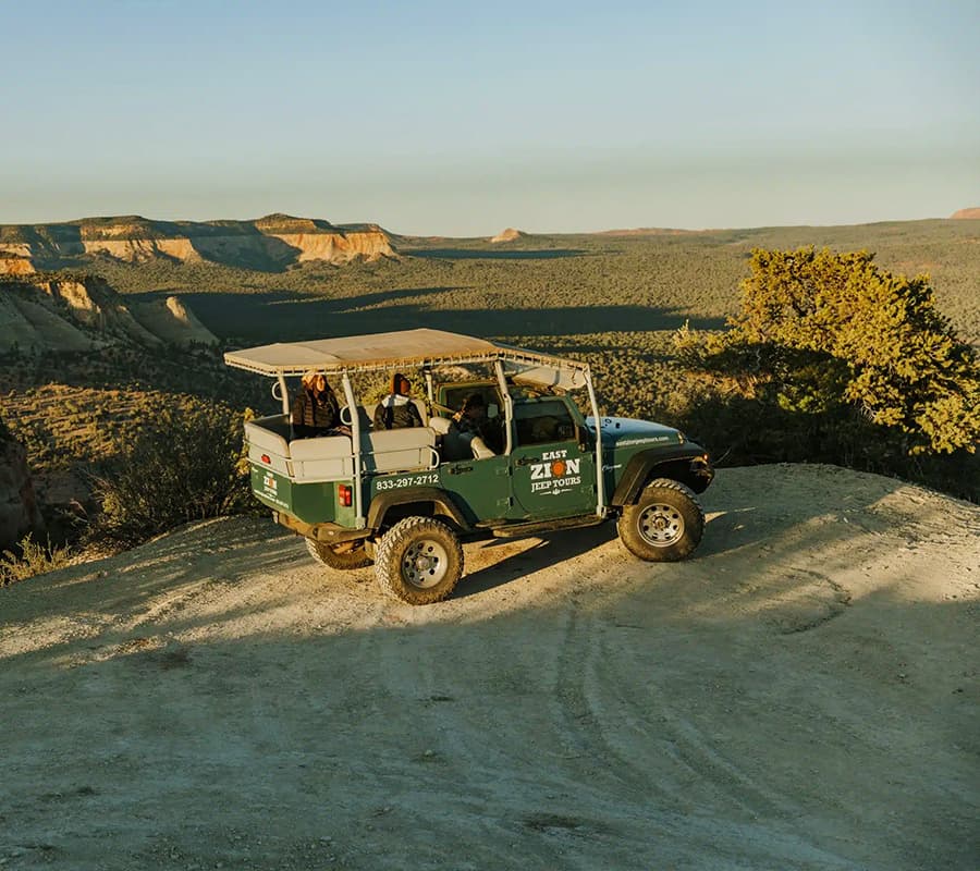 A green jeep with passengers is parked on a scenic overlook, surrounded by vast desert landscapes at sunset.