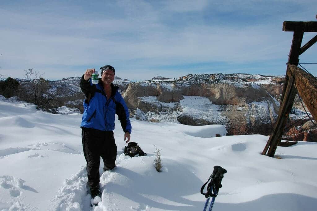 A man in a blue jacket stands in deep snow, smiling and holding a drink against a backdrop of snow-covered cliffs and a bright sky.