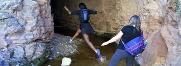 Two hikers navigate through a rocky terrain, one jumping over a small stream.