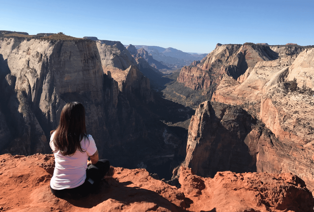 A person sits on a rocky outcrop overlooking a vast, scenic canyon landscape.