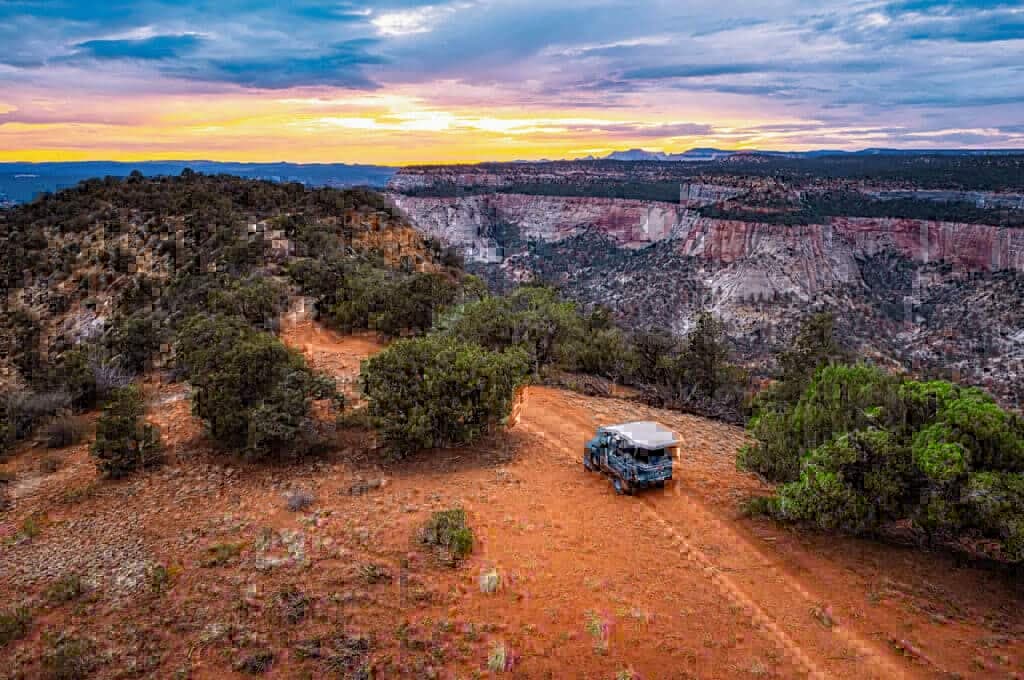 A vehicle parked on a reddish earth path near a canyon at sunset.