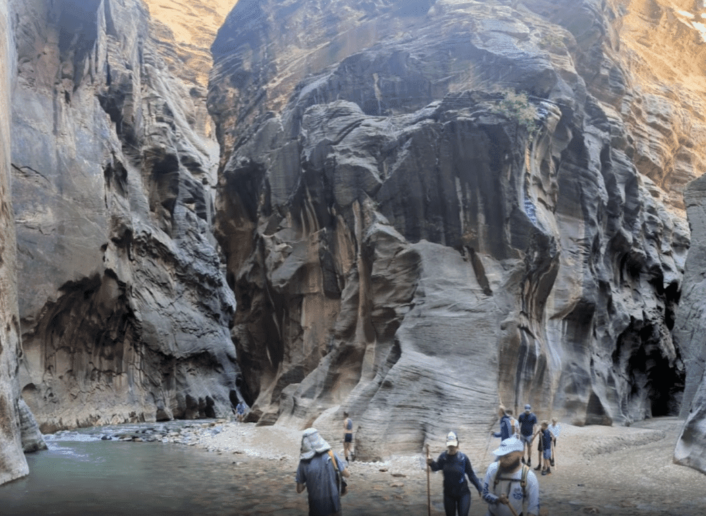 Hikers traverse a narrow canyon with towering rock formations and a stream.