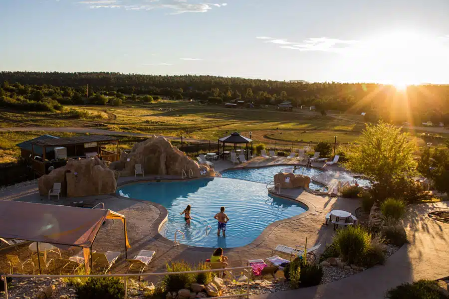 A serene outdoor pool with two people swimming, surrounded by landscape and a sunset.