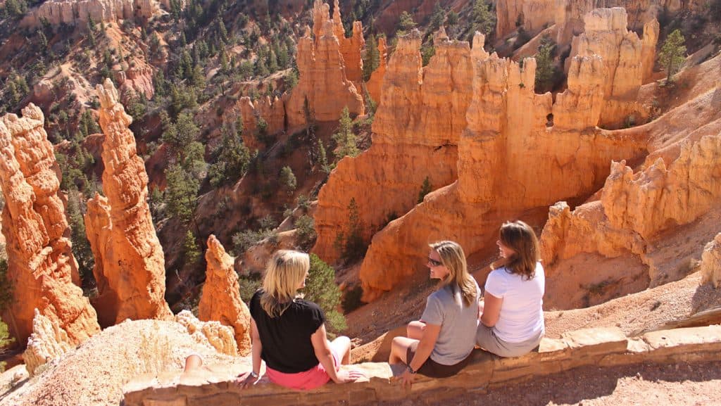Three women sit on a ledge overlooking the orange rock formations of Bryce Canyon.
