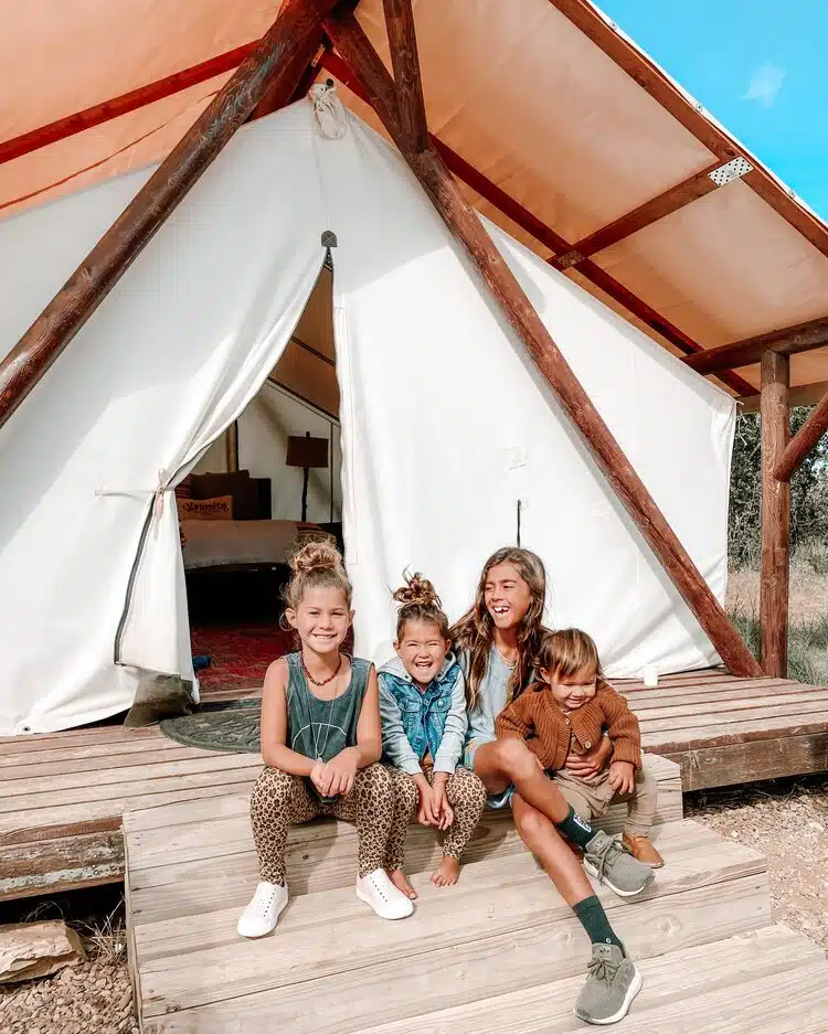 Four children happily sitting on steps in front of a white tent in a natural setting.