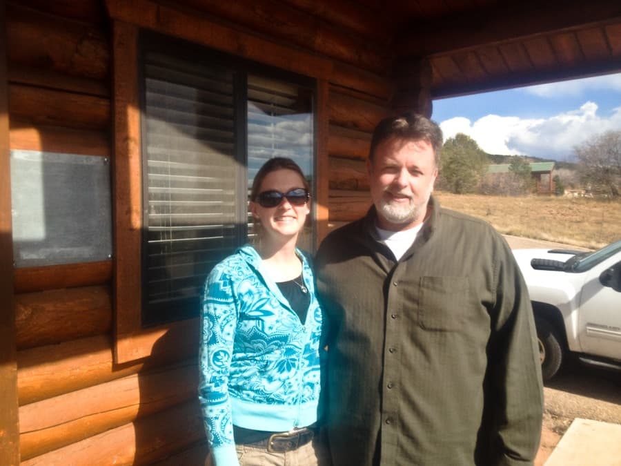 A woman in a blue patterned sweater and a man in a dark shirt pose together in front of a log cabin.