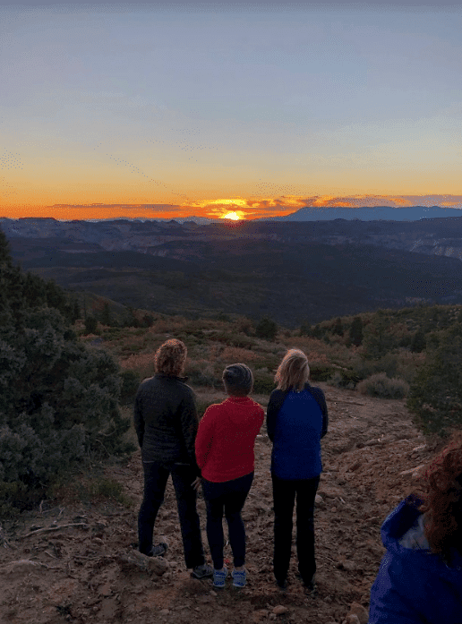 Three people stand on a hillside, gazing at a sunset over a mountainous landscape.