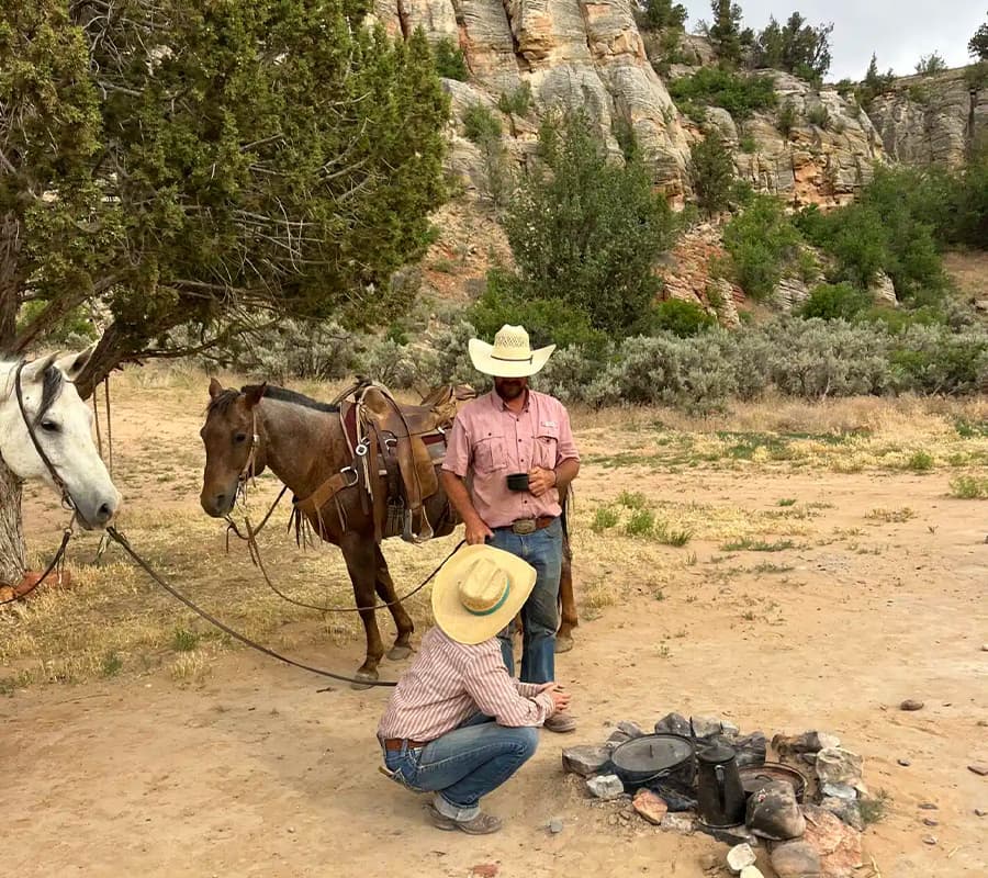 Two cowboys, one standing and one kneeling by a campfire, with horses tethered nearby in a natural landscape.