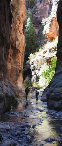 Two hikers navigate a rocky stream in a narrow canyon surrounded by towering sandstone walls.