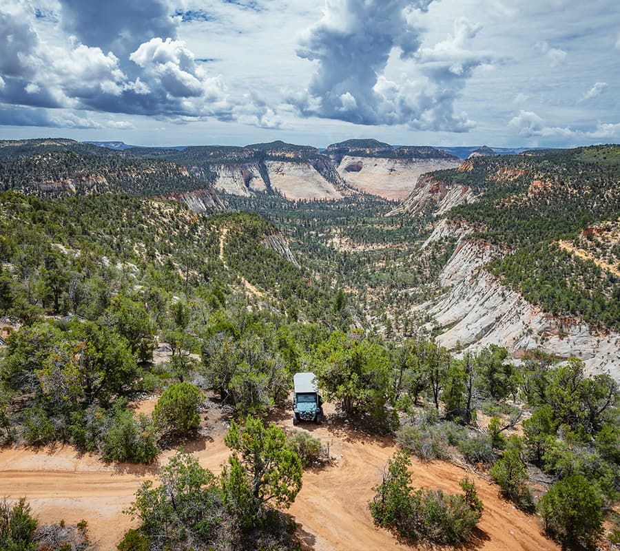 A truck is parked along a winding dirt road overlooking a vast, mountainous valley under a partly cloudy sky.