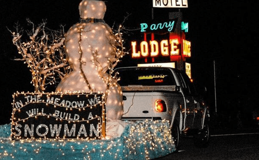 A festive float features a large illuminated snowman and a sign reading "In the meadow we will build a snowman" in front of a motel sign.
