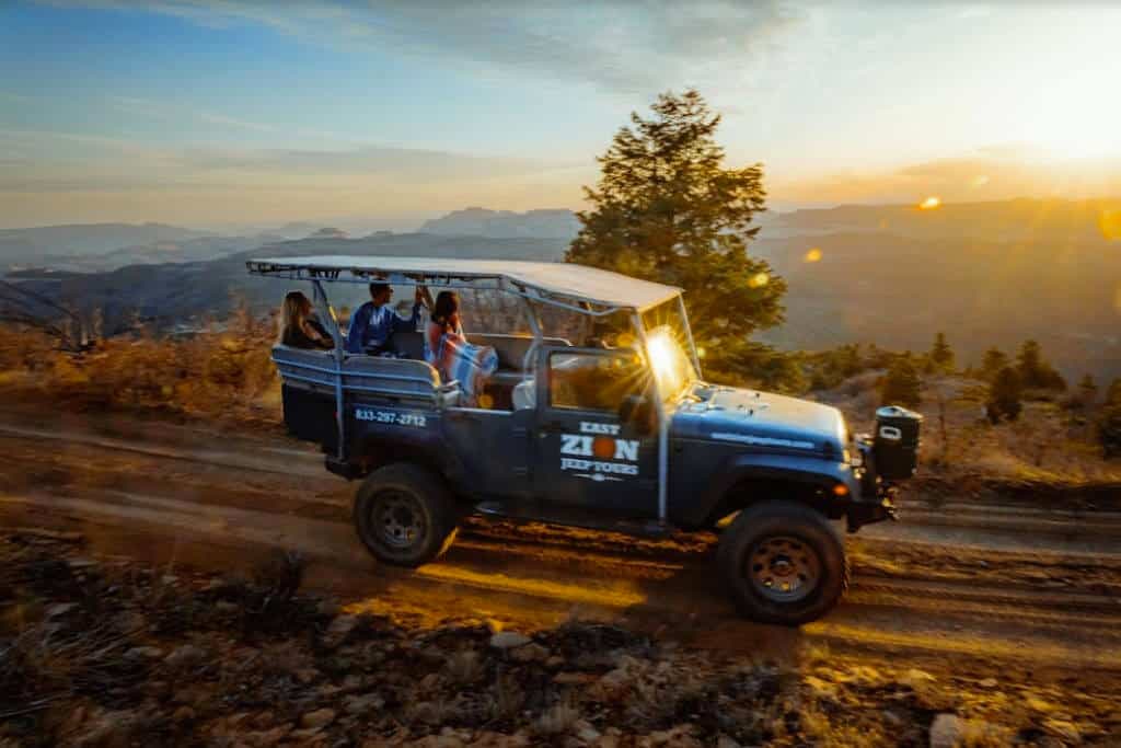 A blue jeep with passengers drives along a rugged dirt road during sunset in a scenic landscape.